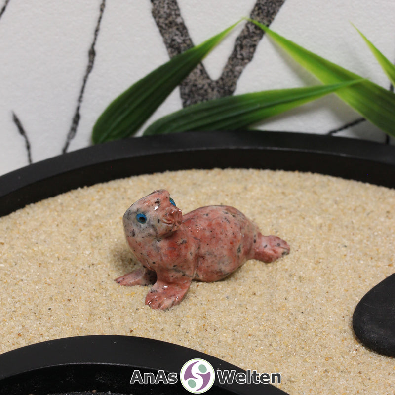 Das Bild zeigt eine Speckstein Robbe klein. Die Specksteinfigur stellt eine Robbe oder einen Seehund dar. Sie liegt auf dem Bauch, ist auf die vorderen Flossen gestützt und hat den Kopf zur Seite gerichtet. Die Augen sind aus einem anderen Material, damit sie besser zur Geltung kommen. Die Details an den Flossen und der Schnauze sind durch Einkerbungen im Stein dargestellt.