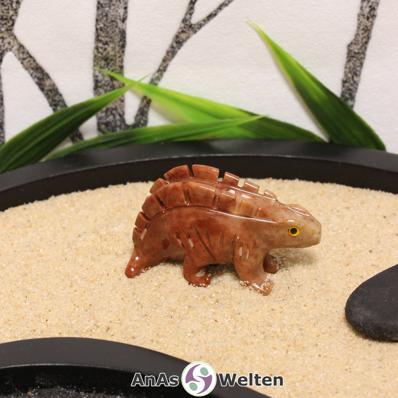 Das Bild zeigt einen Speckstein Stegosaurus klein. Die Specksteinfigur steht auf allen vier Beinen und hat den Kopf nach vorne gerichtet. Der Schwanz ist zur Seite gewinkelt. Die Augen bestehen aus einem anderen Material, damit sie besser zur Geltung kommen, die Rückenplatten sind einzeln herausgearbeitet und die Schuppen und krallen sind durch Einkerbungen im Stein angedeutet.