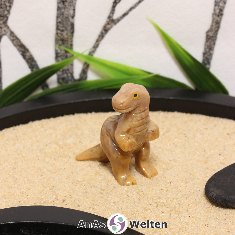 Das Bild zeigt einen Speckstein T-Rex klein. Die Specksteinfigur steht auf den Hinterbeinen und hat den Kopf zur Seite gerichtet. Die Augen bestehen aus einem anderen Material, sodass sie besser zur Geltung kommen, und die Schuppen und Krallen sind durch Einkerbungen im Stein angedeutet.