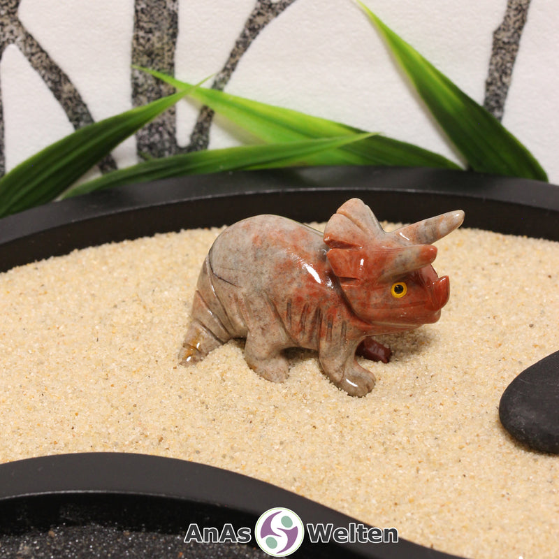 Das Bild zeigt einen Speckstein Stegosaurus klein. Die Specksteinfigur hat den Kopf geradeaus gerichtet und den Schwanz seitlich angewinkelt. Die drei Hörner und der Nackenschild sind aus dem Stein herausgearbeitet, während die Schuppen und Zehen durch Einkerbungen angedeutet sind. Die Augen bestehen aus einem anderen Material, damit sie besser zur Geltung kommen.
