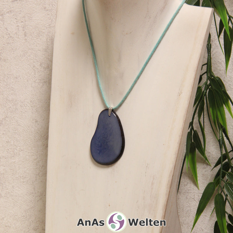 Das Produktbild zeigt die Tagua-Nuss-Schmuck Kette hellblau. Zu sehen ist eine blaue Tagua-Nuss-Scheibe, die mit einer Edelstahlöse an einem türkisen Baumwollband befestigt ist. Die Nuss-Scheibe hat einen dunklen Rand und eine glänzende Oberfläche.