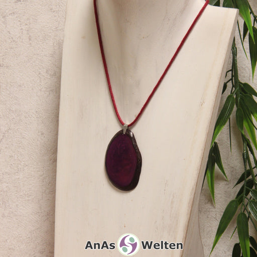 Das Produktbild zeigt die Tagua-Nuss-Schmuck Kette violett. Zu sehen ist eine violette Tagua-Nuss-Scheibe, die mit einer Edelstahlöse an einem roten Baumwollband befestigt ist. Die Nuss-Scheibe hat einen dunklen Rand und eine glänzende Oberfläche.