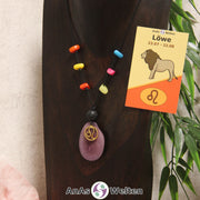 Eine lila Tagua-Nuss Sternzeichen Kette mit bunten Taguasplittern, goldenem Löwe-Symbol und Lavakugel an einer dunklen Büste.