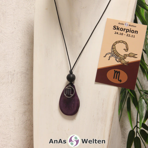 Das Produktbild für die Tagua-Nuss-Schmuck Sternzeichen-Kette Skorpion zeigt eine in violett gefärbte Tagua-Nuss-Scheibe an einem schwarzen Baumwollband, vor der ein silberfarbener Anhänger mit dem Sternzeichensymbol des Skorpions befestigt ist. Die Nuss-Scheibe hat einen dunklen Rand und eine glänzende Oberfläche. Direkt darüber befindet sich eine Lavakugel. Rechts neben der Kette sieht man außerdem die Sternzeichen Skorpion Karte von AnAs Welten.