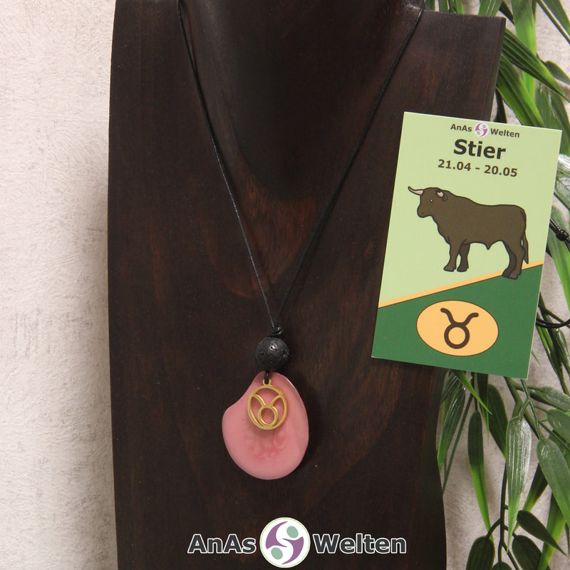 Eine rosa gefärbte Tagua-Nuss Sternzeichen Kette mit goldfarbenem Stier-Symbol und Lavakugel an einer dunklen Büste.