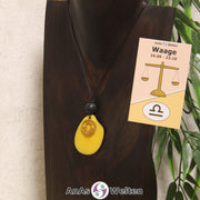 Eine gelb gefärbte Tagua-Nuss Sternzeichen Kette mit goldfarbenem Waage-Symbol und Lavakugel an einer dunklen Büste.