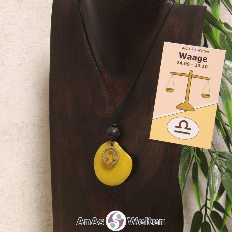 Eine gelb gefärbte Tagua-Nuss Sternzeichen Kette mit silberfarbenem Waage-Symbol und Lavakugel an einer dunklen Büste.