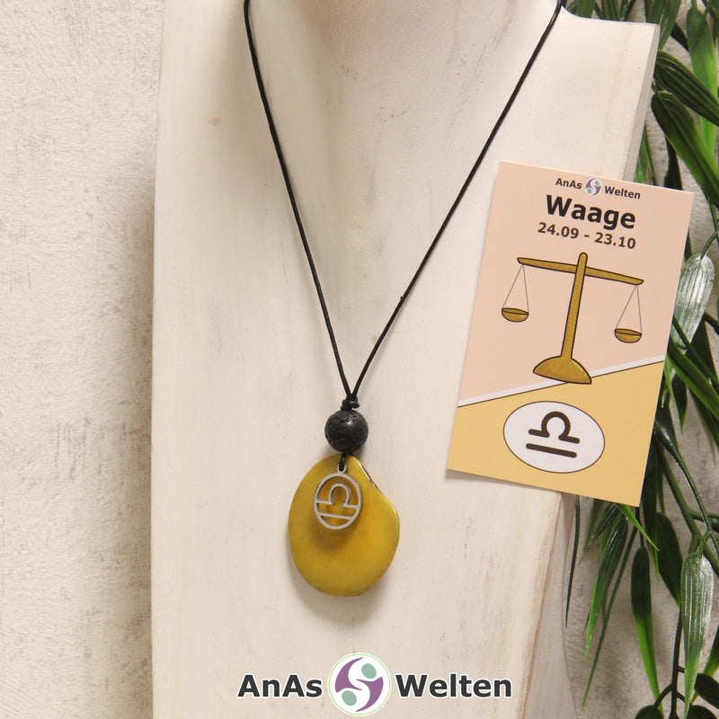 Das Produktbild für die Tagua-Nuss-Schmuck Sternzeichen-Kette Stier zeigt eine gelb gefärbte Tagua-Nuss-Scheibe an einem schwarzen Baumwollband, vor der ein silberfarbener Anhänger mit dem Sternzeichensymbol der Waage befestigt ist. Die Nuss-Scheibe hat einen dunklen Rand und eine glänzende Oberfläche. Direkt darüber befindet sich eine Lavakugel. Rechts neben der Kette sieht man außerdem die Sternzeichen Waage Karte von AnAs Welten.