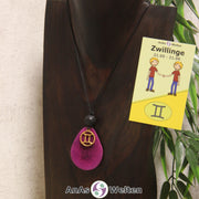 Eine violett gefärbte Tagua-Nuss Sternzeichen Kette mit goldfarbenem Zwillinge-Symbol und Lavakugel an einer dunklen Büste.