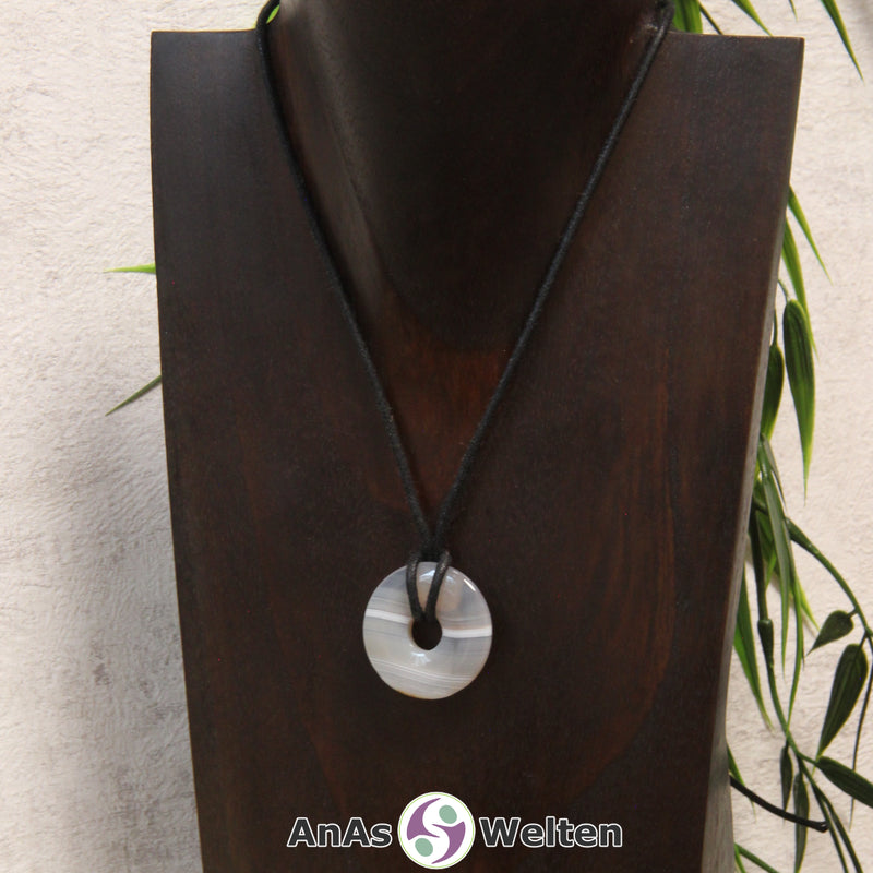 Das Bild zeigt einen Achat Donut an einem Baumwollband. Der Achat Donut ist grau-weiß-graublau gestreift.