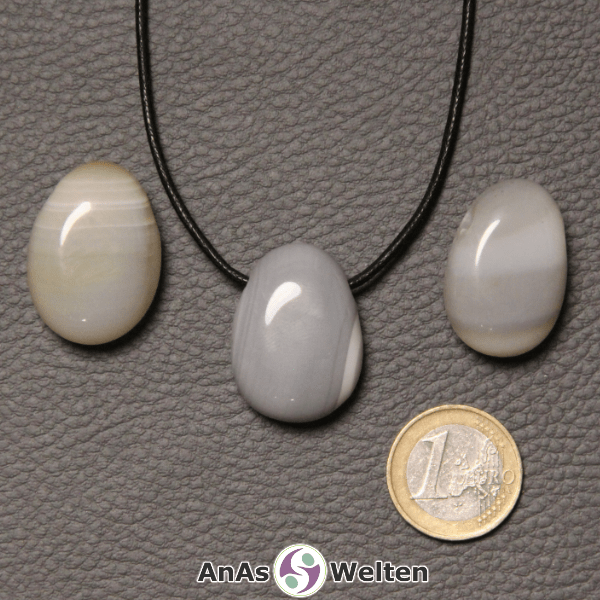 Drei grau-weiße Achat gebohrte Trommelsteine nebeneinander. Einer befindet sich an einem schwarzen gewachsten Baumwollband.