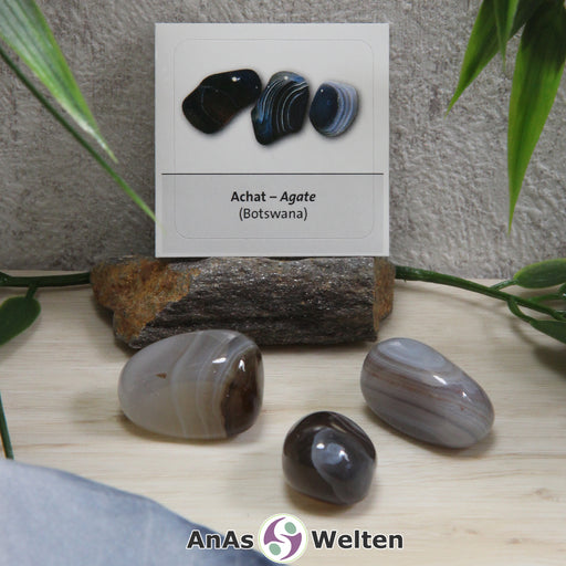 Das Produktbild zeigt den Achat Trommelstein mit Sticker als drei verschiedene Beispielsteine. Die Steine sind grau, grau-braun und braun und weisen dünne helle und dunkle Streifen auf. Im Hintergrund ist der Sticker zu sehen.