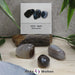 Das Produktbild zeigt den Achat Trommelstein mit Sticker als drei verschiedene Beispielsteine. Die Steine sind grau, grau-braun und braun und weisen dünne helle und dunkle Streifen auf. Im Hintergrund ist der Sticker zu sehen.