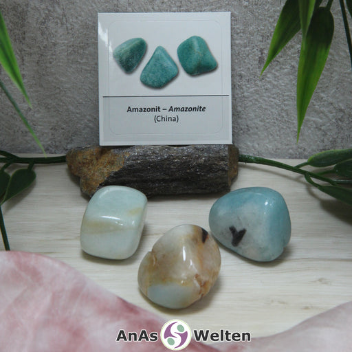 Das Bild zeigt drei Beispiele für den Amazonit Trommelstein mit Sticker. Der Amazonit ist ein hellblauer Stein mit weißen, braunen und/oder weißen Einschlüssen. Im Hintergrund ist der Sticker zu sehen.