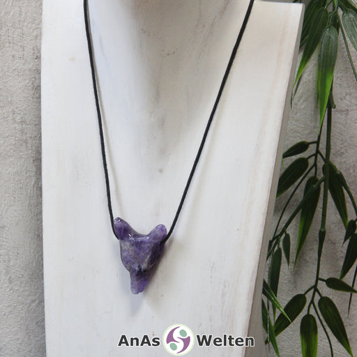 Das Bild zeigt einen Amethyst Wolf Anhänger an einem Baumwollband. Der Edelstein hat die typische Lila-Weiß Farbe, für die der Amethyst bekannt ist. Das Fellmuster und die Nase sind durch kleine Kerben im Edelstein dargestellt. Die Augen sind kleine schwarze Perlen.