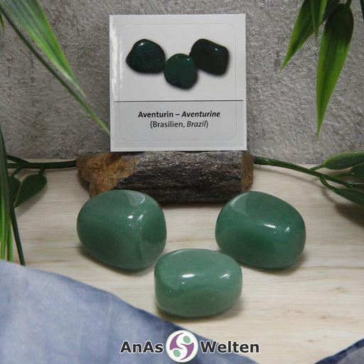 Das Bild zeigt drei Beispielsteine für den Aventurin Trommelstein mit Sticker. Die Steine sind alle in einem dunklen Blaugrün und haben keine erkennbaren Einschlüsse oder Risse. Im Hintergrund ist der Sticker zu sehen.