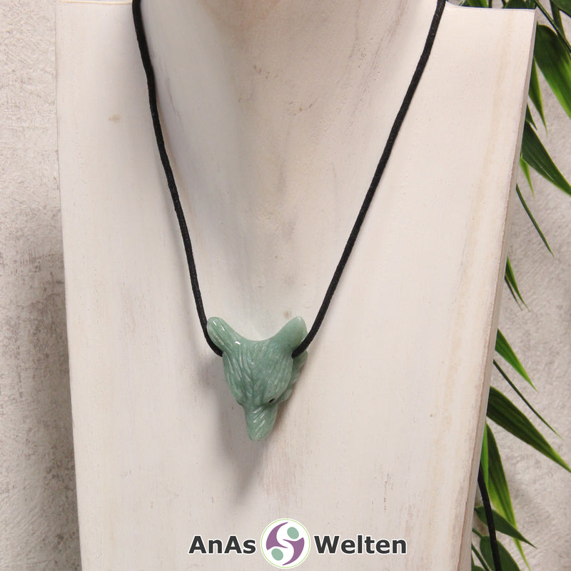 Das Bild zeigt einen Aventurin Wolf Anhänger an einem Baumwollband. Der Edelstein hat eine hellblaue bis hellgrüne Farbe ohne auffällige Musterung. Das Fellmuster und die Nase sind durch kleine Kerben im Edelstein dargestellt. Die Augen sind kleine schwarze Perlen.