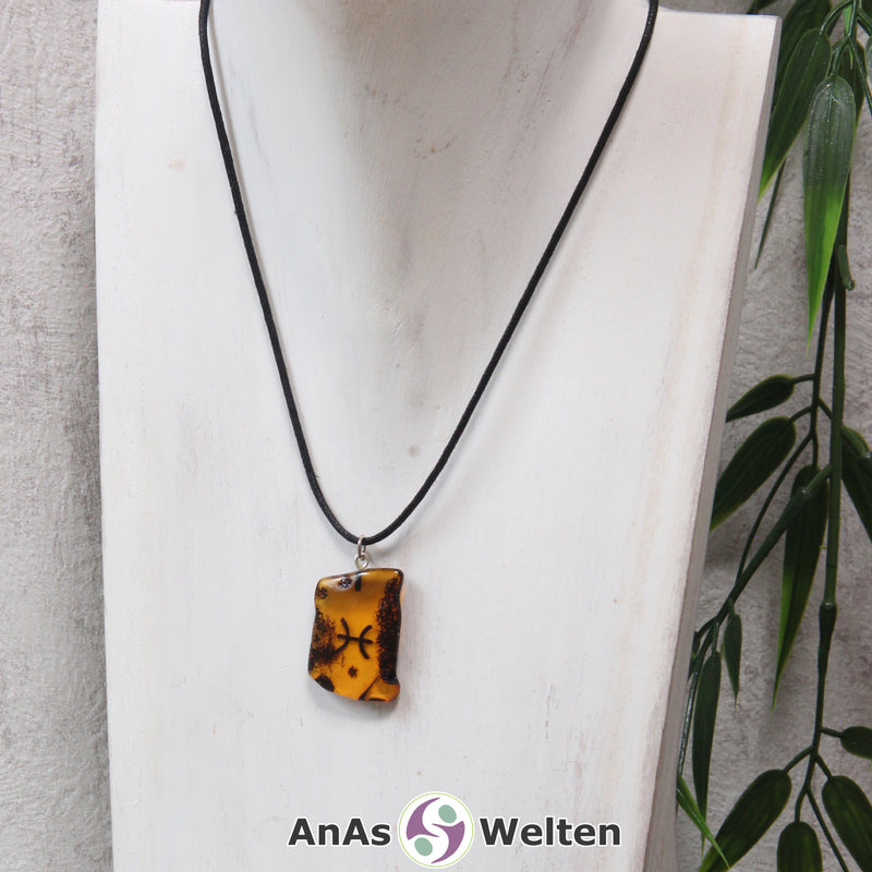 Das Bild zeigt einen Bernstein Anhänger Sternzeichen Fische mit Silberöse an einem Baumwollband. Der Bernstein ist flach, an den ungeschliffenen Kanten poliert und hat die typische honiggoldene Farbe von Bernstein. Da er durchsichtig ist, kann man deutlich das dunkel auf die Rückseite eingravierte Sternzeichensymbol der Fische erkennen.