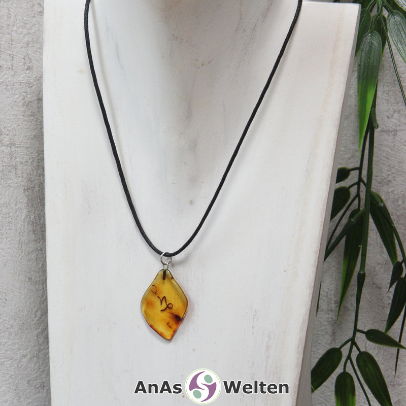 Das Bild zeigt einen Bernstein Anhänger Sternzeichen Steinbock mit Silberöse an einem Baumwollband. Der Bernstein ist flach, an den ungeschliffenen Kanten poliert und hat die typische honiggoldene Farbe von Bernstein. Da er durchsichtig ist, kann man deutlich das dunkel auf die Rückseite eingravierte Sternzeichensymbol des Steinbocks erkennen.