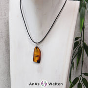 Das Bild zeigt einen Bernstein Anhänger Sternzeichen Wassermann mit Silberöse an einem Baumwollband. Der Bernstein ist flach, an den ungeschliffenen Kanten poliert und hat die typische honiggoldene Farbe von Bernstein. Da er durchsichtig ist, kann man deutlich das dunkel auf die Rückseite eingravierte Sternzeichensymbol des Wassermanns erkennen.