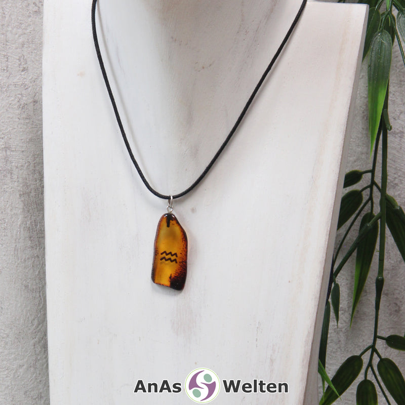 Das Bild zeigt einen Bernstein Anhänger Sternzeichen Wassermann mit Silberöse an einem Baumwollband. Der Bernstein ist flach, an den ungeschliffenen Kanten poliert und hat die typische honiggoldene Farbe von Bernstein. Da er durchsichtig ist, kann man deutlich das dunkel auf die Rückseite eingravierte Sternzeichensymbol des Wassermanns erkennen.