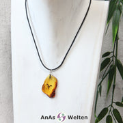 Das Bild zeigt einen Bernstein Anhänger Sternzeichen Widder mit Silberöse an einem Baumwollband. Der Bernstein ist flach, an den ungeschliffenen Kanten poliert und hat die typische honiggoldene Farbe von Bernstein. Da er durchsichtig ist, kann man deutlich das dunkel auf die Rückseite eingravierte Sternzeichensymbol des Widders erkennen.