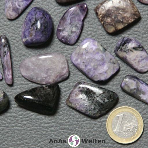 Das Bild zeigt mehrere Beispiele für den Charoit Trommelstein. Die Edelsteine sind meist eine Mischung aus Schwarz und Lila, wobei bei einigen die eine Farbe deutlich überwiegt. Außerdem haben sie oft viele Risse im Inneren, die man in den schwach transparenten Steinen erkennen kann.