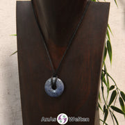 Das Bild zeigt einen Dumortierit Donut an einem Baumwollband. Der Edelstein hat eine dunkelblaue Grundfarbe mit hellblauen bis grau-blauen Flecken.