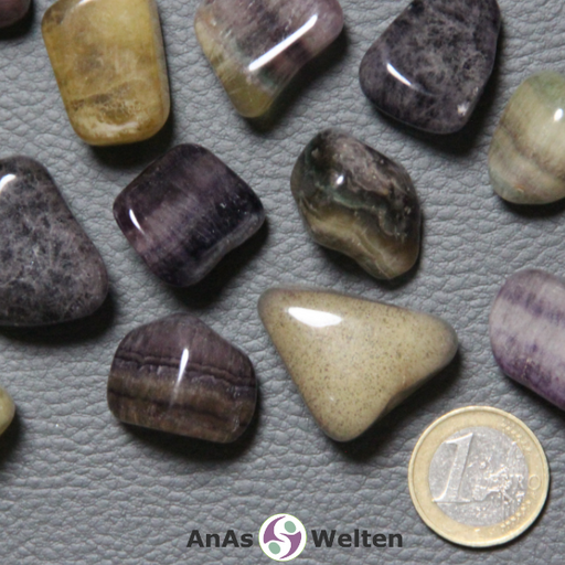 Das Bild zeigt den Fluorit Trommelstein anhand von mehreren Beispielen. Es zeigt gut, wie unterschiedlich der Fluorit aussehen kann. Einige Steine haben ein sandiges Geld, andere sind gelb-lila und andere nur lila. Auch sind einige von ihnen gestreift, während andere relativ einfarbig aussehen.