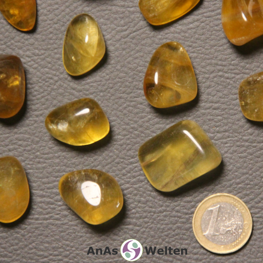 Ein gelber Fluorit Trommelstein wird in mehreren Beispielsteinen dargestellt. Sie haben allesamt eine gelbliche Färbung und sind transparent. Einige von ihnen sind heller, andere dunkler.