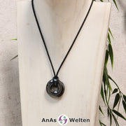 Ein Hämatit Donut als Kette an einem schwarzen gewachsten Baumwollband um eine helle Büste.