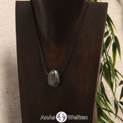 Ein gebohrter Hämatit Trommelstein an einem Baumwollband. Der Edelstein hat eine grau-metallene Oberfläche, die glänzt und spiegelt.