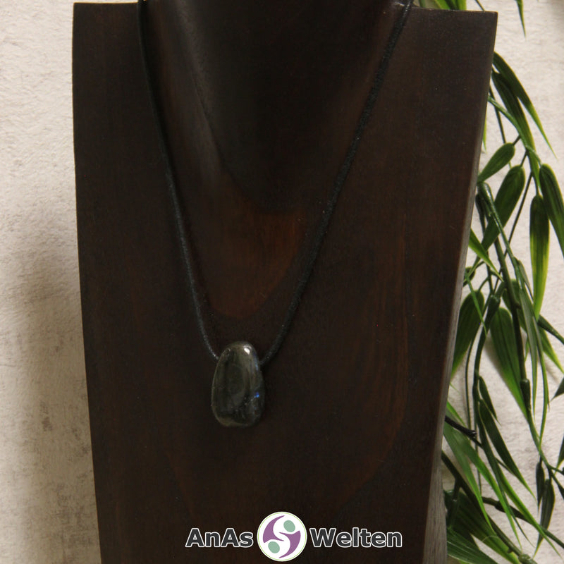 Ein blauschimmernder Labradorit gebohrter Trommelstein an einem schwarzen Baumwollband, der um eine dunkle Büste gehängt wurde.