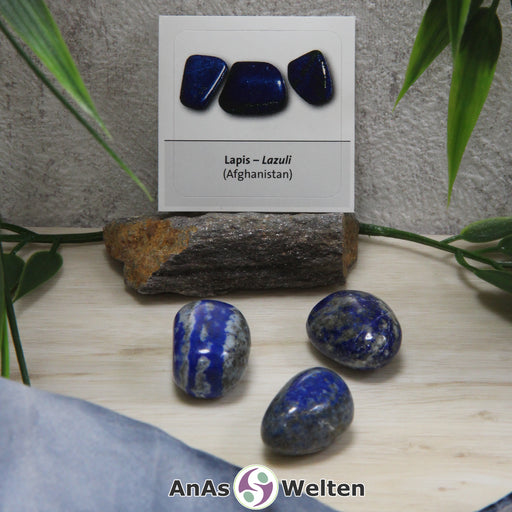 Das Bild zeigt den Lapispazuli Trommelstein mit Sticker anhand von drei Beispielsteinen. Die Steine haben einen tief dunkelblauen Farbton mit einer grauen Musterung. Im Hintergrund ist der Sticker zu sehen.