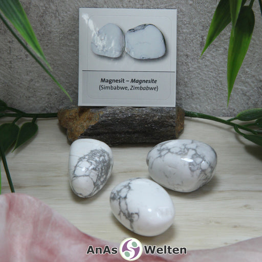 Das Bild zeigt einen Magnesit Trommelstein mit Sticker anhand von drei Beispielsteinen. Die Steine erinnern optisch stark an Marmor, sind also weiß und mit grauen Linien durchzogen. Im Hintergrund ist der Sticker zu sehen.