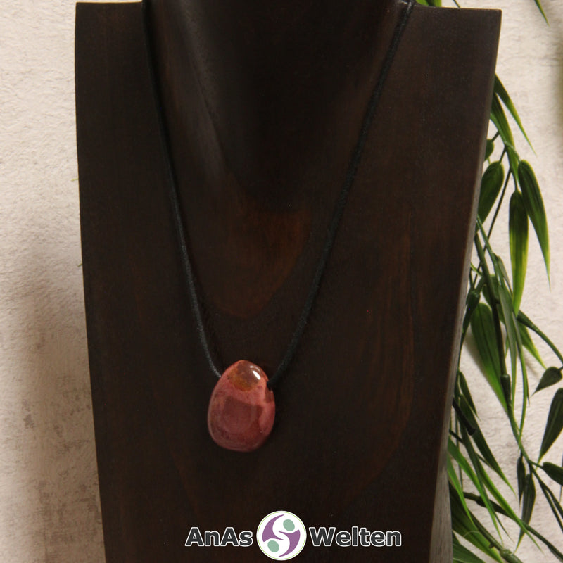 Ein gebohrter Mookait Trommelstein an einem Baumwollband. Der Edelstein hat eine dunkelrote Farbe mit einer hellroten Musterung. Mookait kann aber auch andere Farben wie z. B. ein warmes Gelb haben.