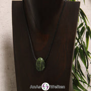 Ein gebohrter Nephrit Jade Trommelstein an einem Baumwollband. Der Edelstein hat eine Mischung aus dunkel- und Hellgrün. An einigen Stellen ist er von schwarzen Punkten durchzogen.