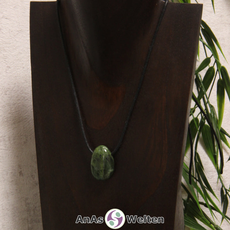 Ein gebohrter Nephrit Jade Trommelstein an einem Baumwollband. Der Edelstein hat eine Mischung aus dunkel- und Hellgrün. An einigen Stellen ist er von schwarzen Punkten durchzogen.
