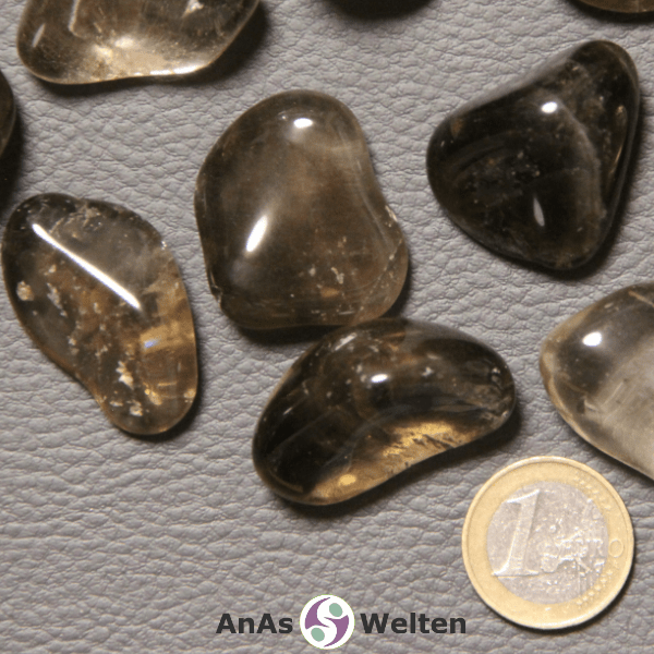 Das Produktbild zeigt einen Rauchquarz Trommelstein anhand mehrerer Beispielsteine. Die Edelsteine sind grau-braun und größtenteils durchsichtig, wodurch man die Einschlüsse und Risse in ihrem Inneren gut sehen kann.