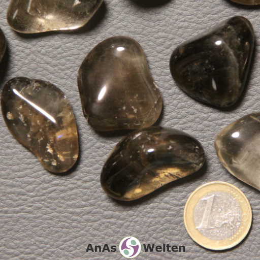 Das Produktbild zeigt einen Rauchquarz Trommelstein anhand mehrerer Beispielsteine. Die Edelsteine sind grau-braun und größtenteils durchsichtig, wodurch man die Einschlüsse und Risse in ihrem Inneren gut sehen kann.