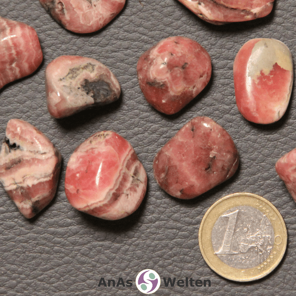 Das Bild zeigt einen Rhodochrosit Trommelstein anhand mehrerer Beispielsteine. Die Edelsteine haben allesamt eine Grundfarbe aus Lachs und Beige. Außerdem haben die meisten von ihnen grau-schwarze Einschlüsse.