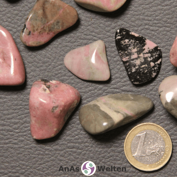 Das Bild zeigt einen Rhodonit Trommelstein anhand mehrerer Beispielsteine. Die Edelsteine sehen teilweise sehr verschieden aus, haben aber fast alle einen lachsfarbenen Grundton. Viele haben außerdem einen gewissen Beigeanteil, während einer hauptsächlich graubraun ist und ein anderer lachs und schwarz.