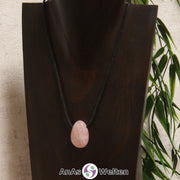 Ein gebohrter Rosenquarz Trommelstein an einem Baumwollband. Der Edelstein ist rosa. In seinem Inneren sind mehrere helle Risse zu erkennen.