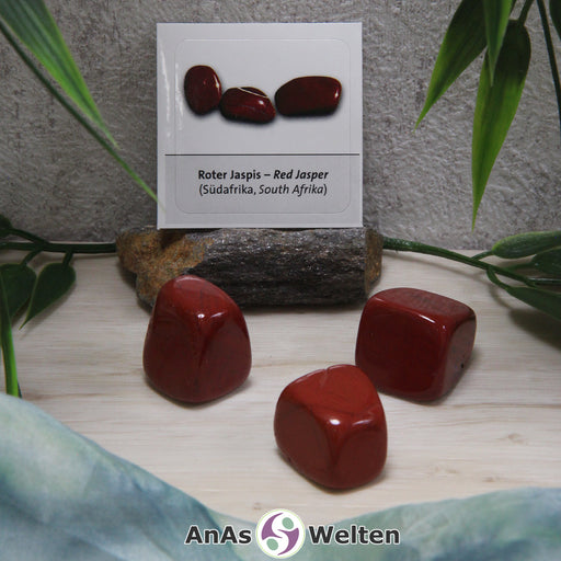 Ein roter Jaspis Trommelstein mit Sticker wird als drei Beispielsteine dargestellt. Die Edelsteine haben allesamt einen kräftigen roten Farbton. Außerdem haben sie gelegentliche dunkle Einschlüsse. Im Hintergrund ist der Sticker zu sehen.