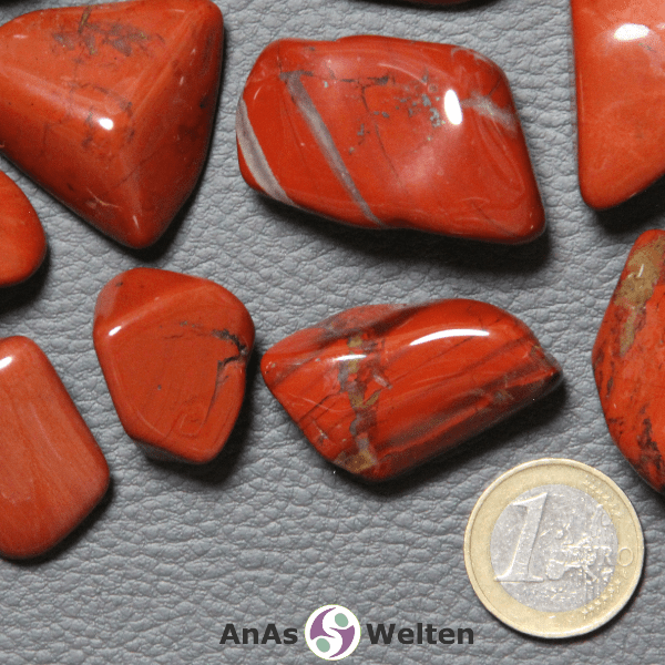 Auf dem Bild ist ein roter Jaspis Trommelstein anhand mehrerer Beispielsteine dargestellt. Die Edelsteine haben eine kräftige rote Grundfarbe sowie mehrere graue und/oder braune Einschlüsse.