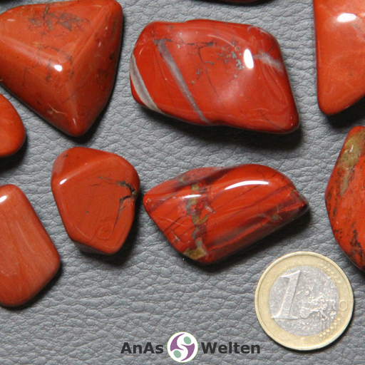 Auf dem Bild ist ein roter Jaspis Trommelstein anhand mehrerer Beispielsteine dargestellt. Die Edelsteine haben eine kräftige rote Grundfarbe sowie mehrere graue und/oder braune Einschlüsse.