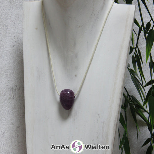 Ein gebohrter Rubin Trommelstein an einem Baumwollband. Der Edelstein hat eine dunkelrote fast lila Farbe. In seinem Inneren sind mehrere helle Einschlüsse zu erkennen.