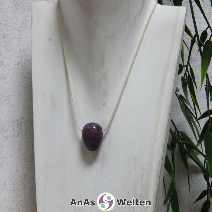 Ein gebohrter Rubin Trommelstein an einem Baumwollband. Der Edelstein hat eine dunkelrote fast lila Farbe. In seinem Inneren sind mehrere helle Einschlüsse zu erkennen.