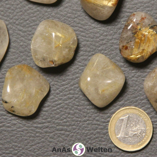 Das Bild zeigt einen Rutilquarz Trommelstein anhand mehrerer Beispielsteine. Die Edelsteine haben eine weiß-gelbliche Grundfarbe und sind halbdurchsichtig. In ihrem Inneren kann man mehrere Risse erkennen, die Golden schimmern.