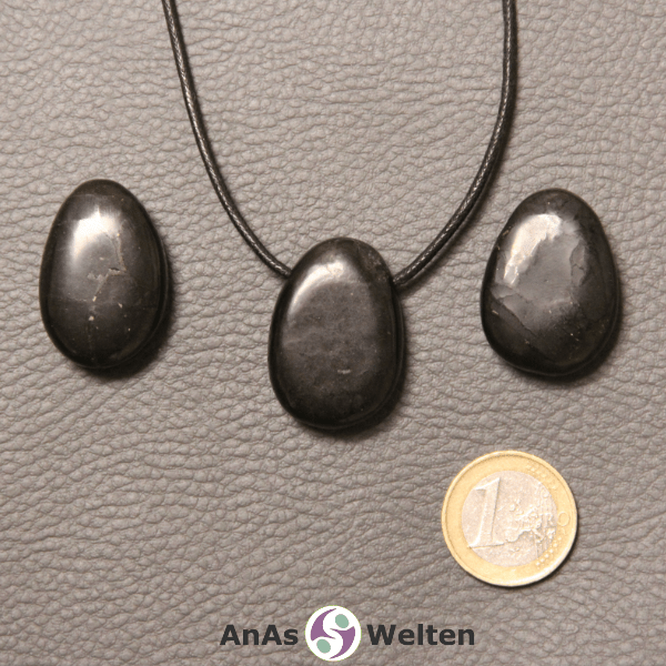 Drei schwarze Schungit gebohrte Trommelsteine nebeneinander. Einer befindet sich an einem schwarzen gewachsten Baumwollband.